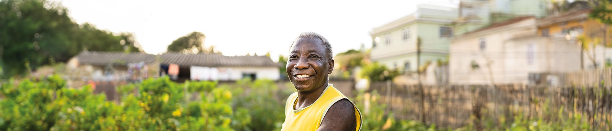 Smiling older person standing amongst crops, holding a wooden gardening tool